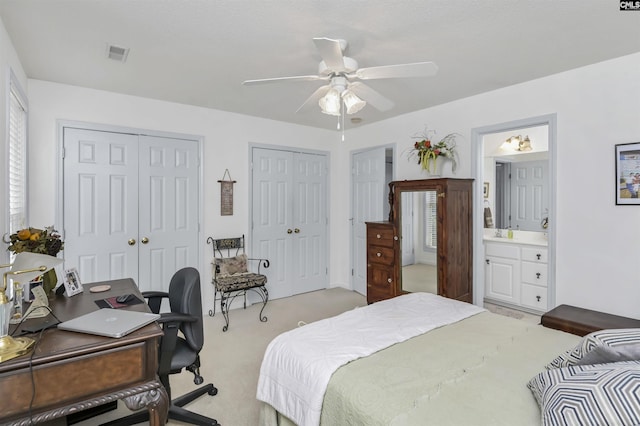 bedroom with ceiling fan, ensuite bathroom, light colored carpet, visible vents, and multiple closets