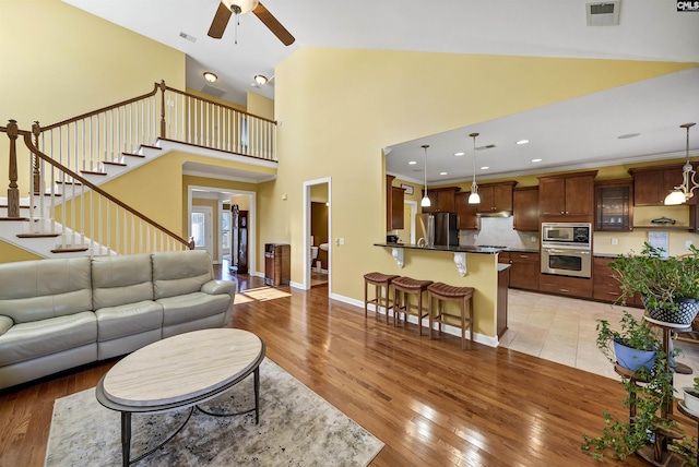 living room featuring visible vents, stairway, light wood-style flooring, baseboards, and ceiling fan with notable chandelier