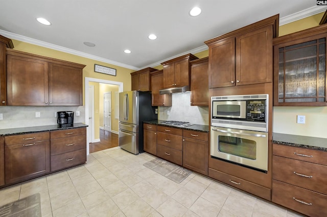 kitchen featuring dark stone countertops, stainless steel appliances, crown molding, under cabinet range hood, and backsplash