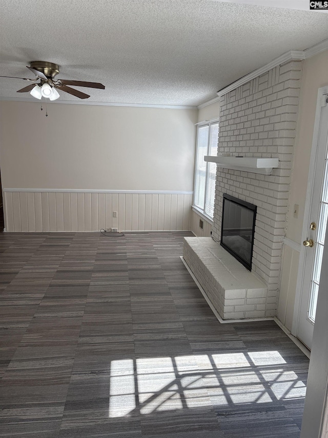 unfurnished living room with a brick fireplace, ceiling fan, ornamental molding, wainscoting, and a textured ceiling