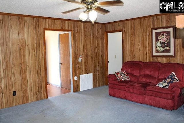 carpeted living room with ornamental molding, visible vents, ceiling fan, and wooden walls