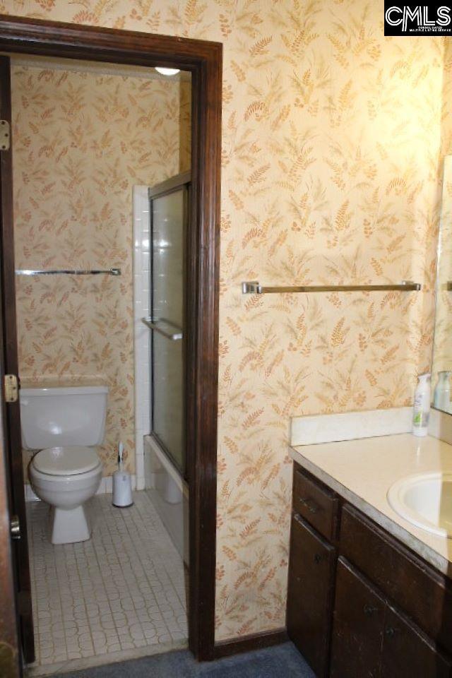 bathroom with toilet, wallpapered walls, shower / bath combination with glass door, and vanity