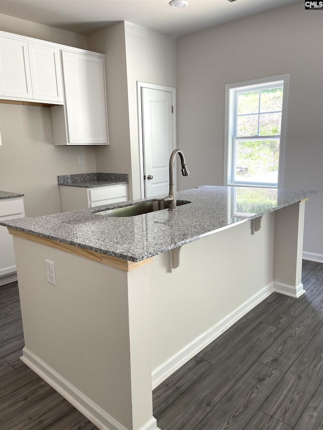 kitchen with light stone counters, a center island with sink, white cabinets, a sink, and baseboards