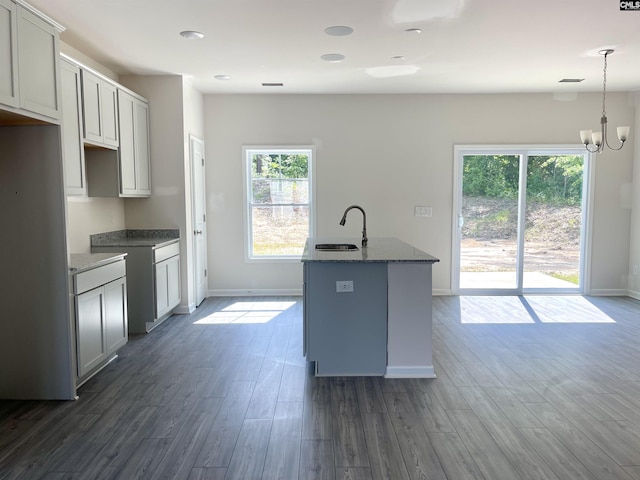 kitchen with dark stone counters, dark wood-style floors, a sink, a kitchen island with sink, and a notable chandelier