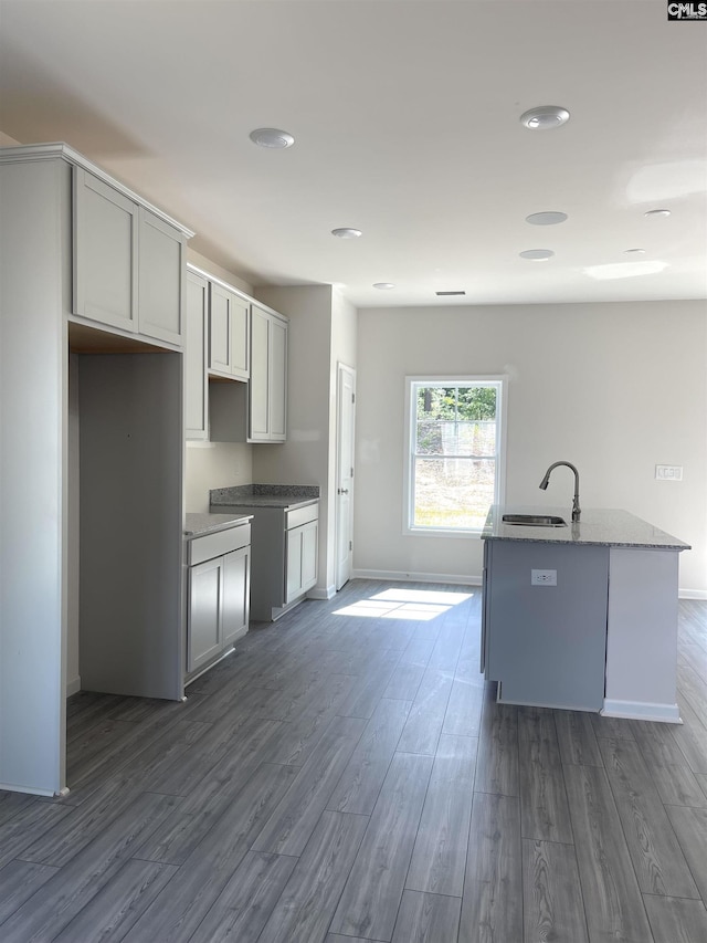 kitchen featuring stone counters, a sink, baseboards, dark wood finished floors, and a center island with sink