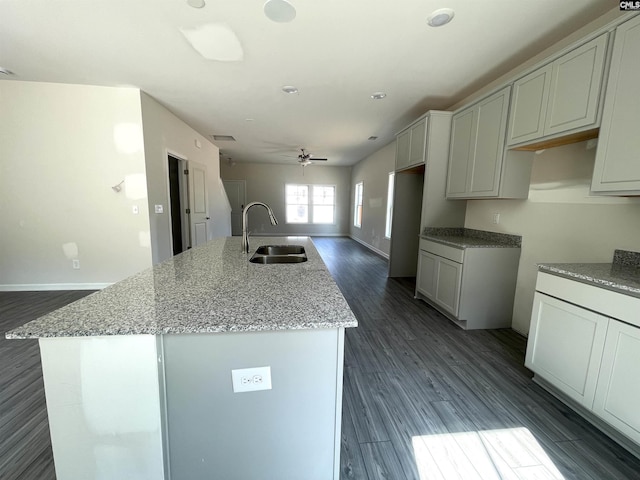 kitchen featuring ceiling fan, dark wood-style flooring, a sink, light stone countertops, and an island with sink