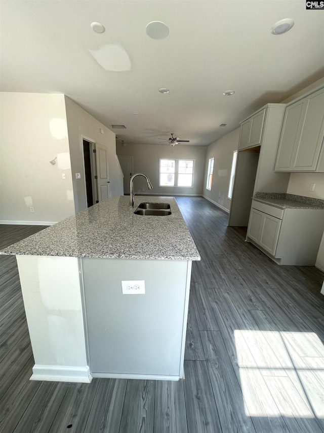 kitchen featuring dark wood finished floors, a sink, an island with sink, light stone countertops, and baseboards