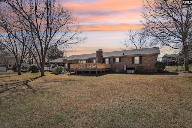 back of property at dusk with a deck, brick siding, crawl space, a lawn, and a chimney