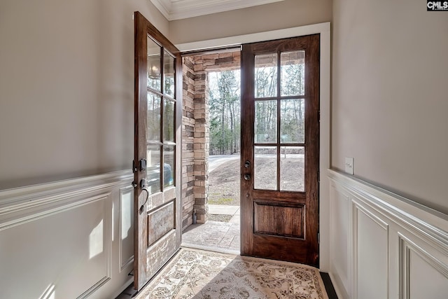 doorway to outside with ornamental molding, a decorative wall, stone tile flooring, and wainscoting