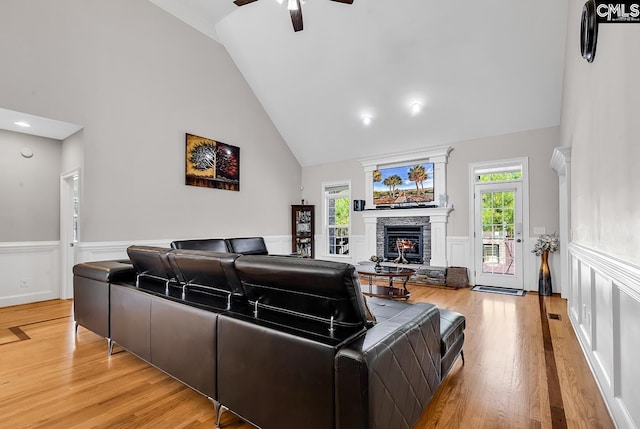 living room with a stone fireplace, wainscoting, high vaulted ceiling, and light wood-type flooring