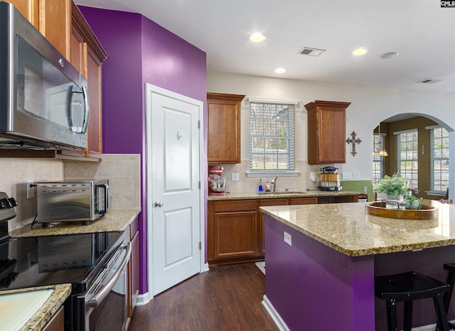 kitchen with a sink, dark wood-style floors, stainless steel appliances, arched walkways, and light stone countertops