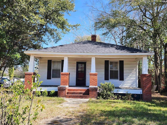 bungalow-style house with covered porch, a chimney, roof with shingles, and a front yard