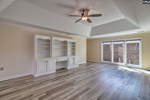 unfurnished living room featuring a tray ceiling, baseboards, built in desk, and light wood finished floors