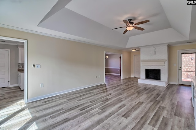 unfurnished living room featuring a raised ceiling, light wood-style floors, baseboards, a brick fireplace, and ceiling fan