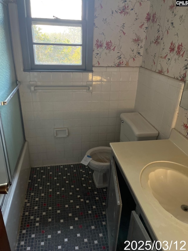 bathroom with toilet, combined bath / shower with glass door, wainscoting, wallpapered walls, and vanity