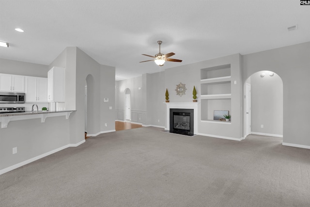 unfurnished living room featuring built in shelves, ceiling fan, baseboards, a fireplace with flush hearth, and arched walkways