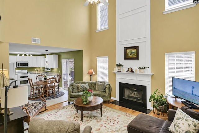 living room featuring visible vents, ceiling fan, a towering ceiling, light wood-type flooring, and a large fireplace