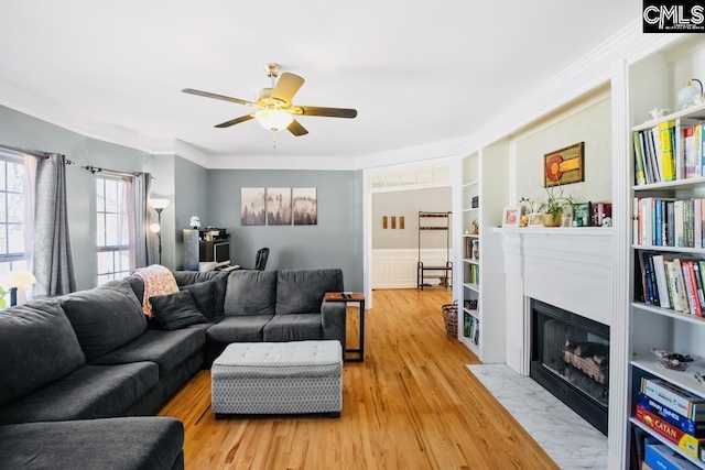 living area featuring built in shelves, a fireplace with flush hearth, crown molding, light wood finished floors, and ceiling fan