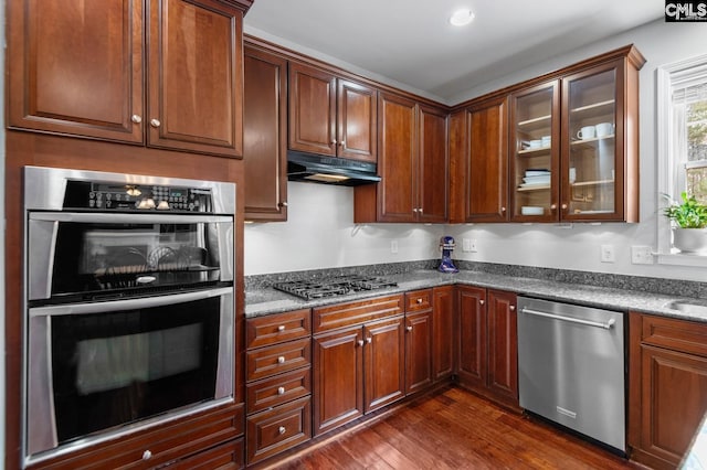 kitchen featuring glass insert cabinets, under cabinet range hood, dark wood finished floors, dark stone counters, and appliances with stainless steel finishes