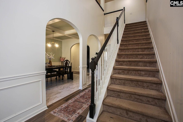 stairs with beamed ceiling, coffered ceiling, wood finished floors, arched walkways, and a chandelier