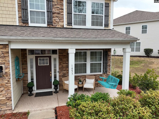 entrance to property featuring stone siding, covered porch, and roof with shingles