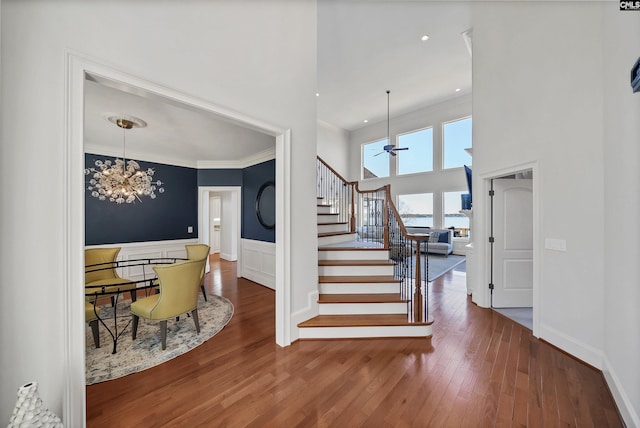 stairs with crown molding, hardwood / wood-style floors, wainscoting, ceiling fan with notable chandelier, and a towering ceiling