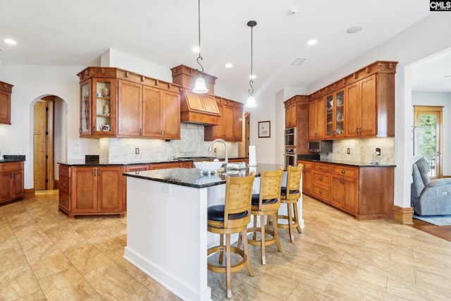 kitchen featuring glass insert cabinets, arched walkways, custom exhaust hood, and a center island with sink