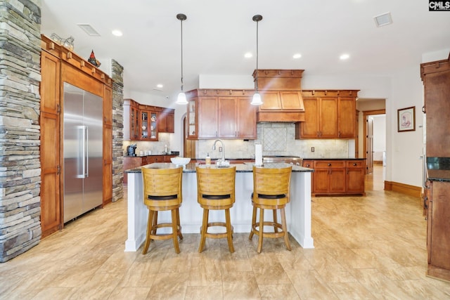 kitchen with visible vents, premium range hood, a kitchen island with sink, built in fridge, and backsplash