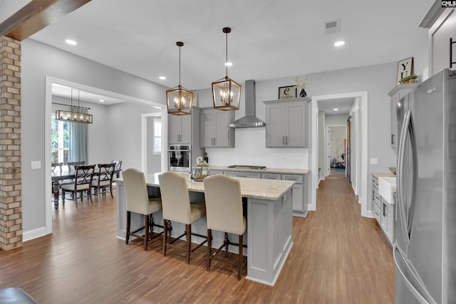 kitchen with visible vents, wood finished floors, stainless steel appliances, an inviting chandelier, and wall chimney exhaust hood