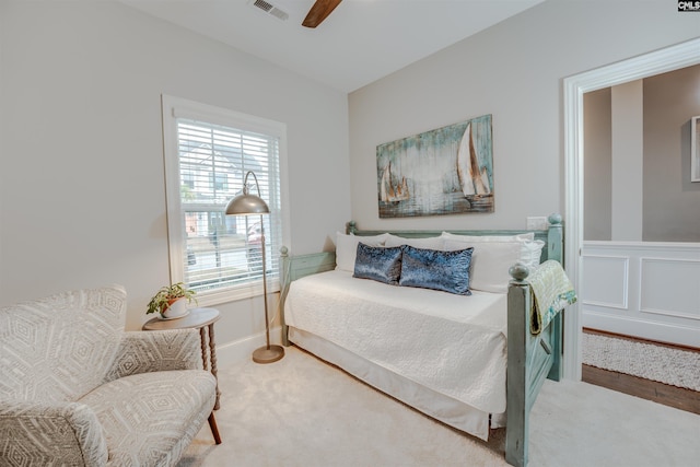 carpeted bedroom featuring ceiling fan, visible vents, a wainscoted wall, and a decorative wall