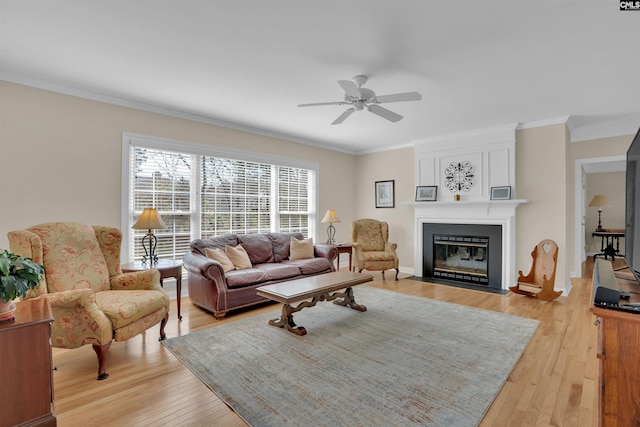 living room featuring light wood-type flooring, a fireplace, crown molding, baseboards, and ceiling fan