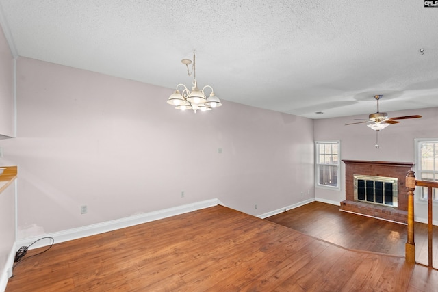 unfurnished living room featuring wood finished floors, baseboards, a textured ceiling, a brick fireplace, and ceiling fan with notable chandelier