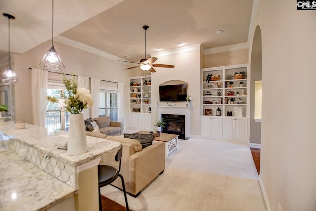 carpeted living room featuring a glass covered fireplace, built in shelves, ceiling fan, and ornamental molding