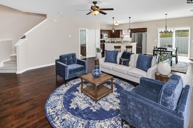 living area with stairway, ceiling fan with notable chandelier, dark wood-style flooring, and baseboards