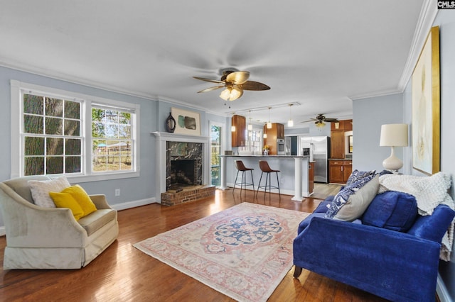 living area featuring a ceiling fan, a fireplace, wood finished floors, baseboards, and ornamental molding