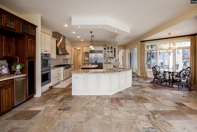 kitchen featuring a chandelier, wall chimney exhaust hood, appliances with stainless steel finishes, glass insert cabinets, and a sink