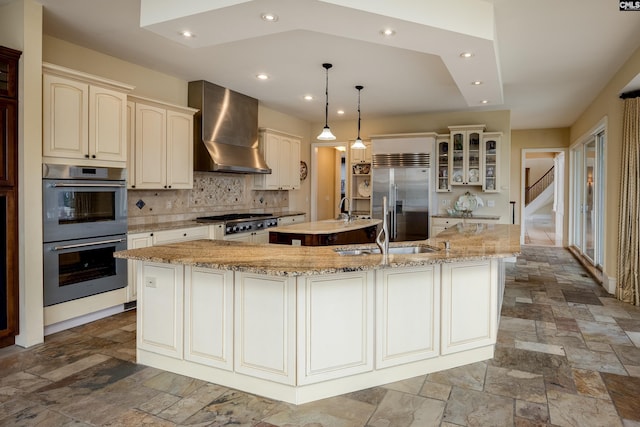 kitchen featuring wall chimney exhaust hood, stone tile flooring, appliances with stainless steel finishes, a spacious island, and cream cabinets