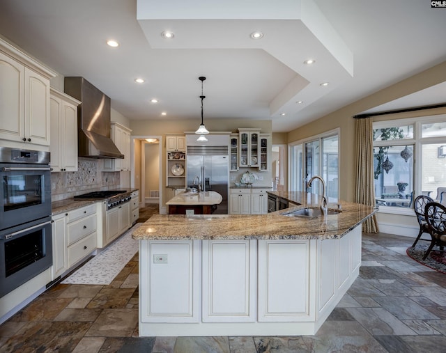 kitchen featuring a large island with sink, stone tile floors, stainless steel appliances, a sink, and wall chimney range hood
