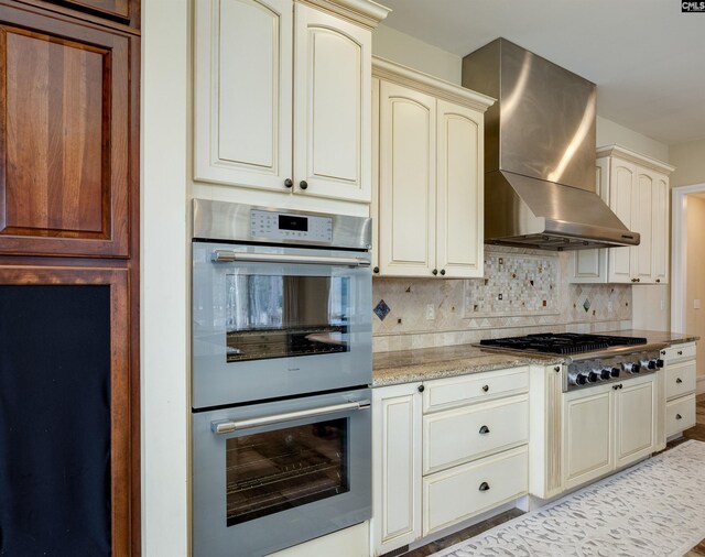kitchen featuring light stone countertops, cream cabinets, stainless steel appliances, wall chimney range hood, and decorative backsplash
