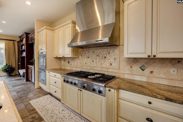 kitchen featuring cream cabinets, stone tile flooring, stainless steel appliances, and wall chimney exhaust hood