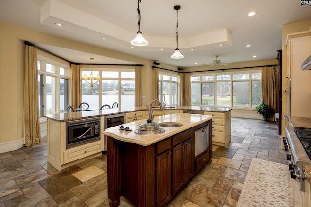 kitchen with a kitchen island with sink, a sink, built in microwave, a tray ceiling, and stone tile floors
