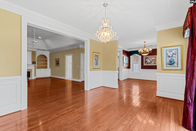 unfurnished living room with hardwood / wood-style floors, ornamental molding, ceiling fan with notable chandelier, a wainscoted wall, and a fireplace