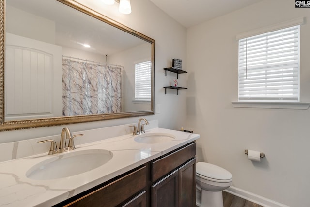 full bath with toilet, a sink, double vanity, and baseboards