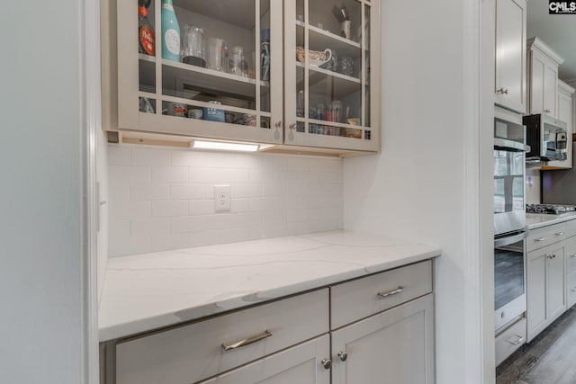 kitchen with tasteful backsplash, stainless steel appliances, dark wood-type flooring, light stone countertops, and glass insert cabinets
