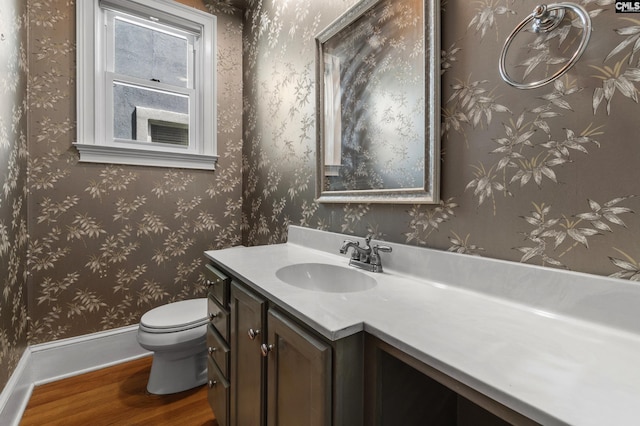 half bathroom with baseboards, toilet, vanity, wood finished floors, and wallpapered walls