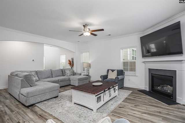 living room with arched walkways, light wood-style flooring, ornamental molding, ceiling fan, and a fireplace with flush hearth