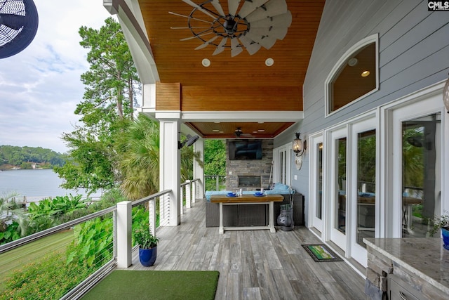 wooden deck with a water view, ceiling fan, and an outdoor stone fireplace
