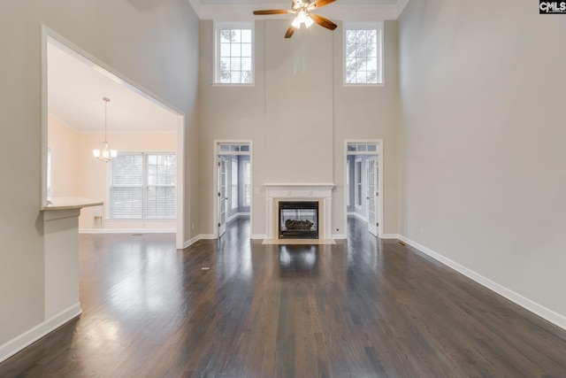 unfurnished living room featuring baseboards, a high ceiling, dark wood-type flooring, a fireplace with flush hearth, and ceiling fan with notable chandelier