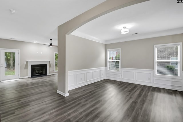 unfurnished living room with ceiling fan, a fireplace, arched walkways, a wainscoted wall, and dark wood-type flooring