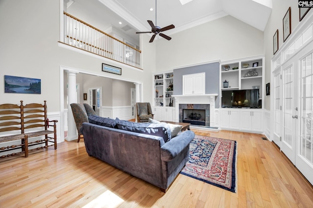 living room featuring ceiling fan, a towering ceiling, a fireplace, light wood-type flooring, and built in shelves